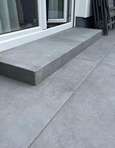 Graue Terrasse mit breiter Betonstufe vor einer weißen Tür, angelehnte Gartenstühle stehen rechts an der Wand.