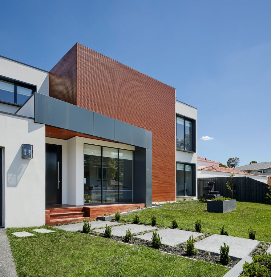 Modernes Flachdachhaus mit weißer Fassade, roter Holzverkleidung und großem Fenster, davor ein gepflegter Garten mit Weg.