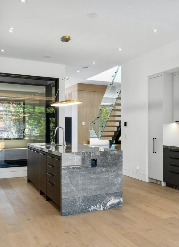 Moderne Küche mit Kochinsel aus grauem Naturstein, hellem Holzfußboden und freitragender Treppe mit Glasgeländer im Hintergrund.