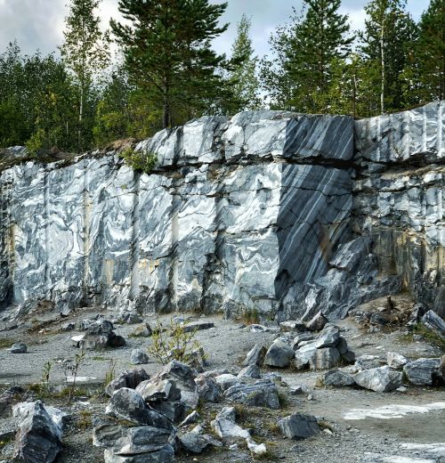 Offener Steinbruch mit grau-weiß gemasertem Gestein, teils bewachsen, locker verstreute Steinbrocken im Vordergrund.