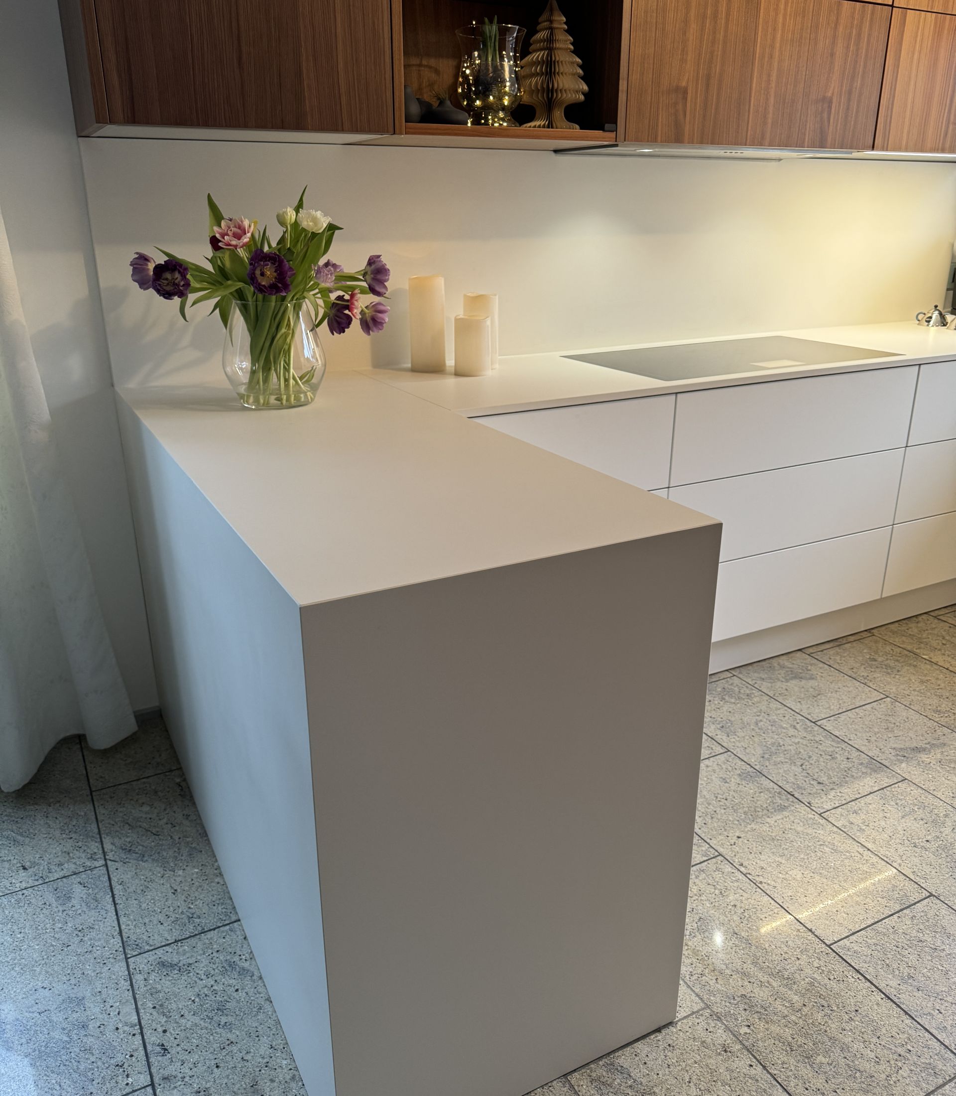 Moderne Küche mit weißer Kochinsel, Tulpenvase, Holzoberschränken und Kerzen auf glänzendem Steinboden.